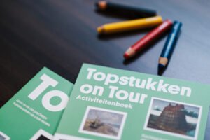 Op een donker houten tafelblad liggen twee magazines. De voorkanten zijn groen. Op de voorkant staat Topstukken on Tour. Naast de tijdschriften liggen dikke potloden in de kleuren: zwart, geel, rood en blauw.