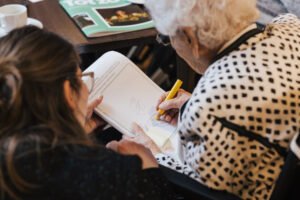 Een ouderen vrouw houdt een tekenboek vast en maakt een tekening met een geel potlood. Een vrouw naast haar houdt het tekenboek vast.